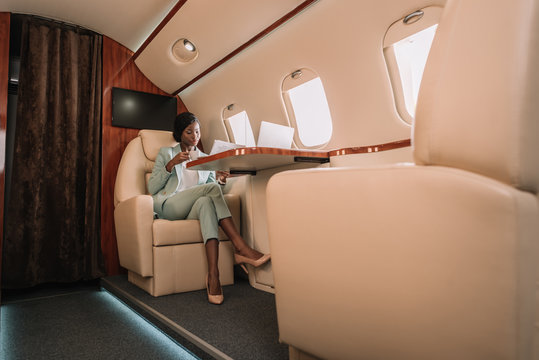 confident african american businesswoman drinking coffee and reading newspaper in private jet