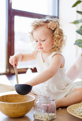 Obraz premium A little girl is sitting on a table in a bright Scandinavian-style kitchen. Playing with dishes, cereals, beans. The concept of children's development Montessori.