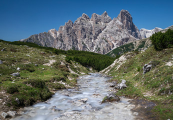 Fototapeta na wymiar River between meadow with mountain range in the background at Three Peaks nature reserve, blue sky during summer, South Tyrol Italy.