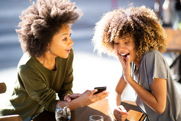 two cheerful young african american women sitting at outdoor cafe looking at cellphone
