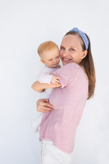 Cheerful positive young mom holding and embracing baby, looking an camera, smiling. Young woman with little daughter isolated over outdoor white wall. Child care concept