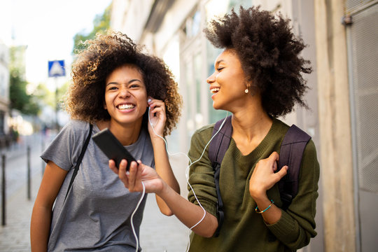 two happy female black friends walking in city with mobile phone listening to music with earphones