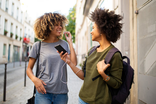 happy female black friends walking in city with mobile phone listening to music with earphones - Powered by Adobe