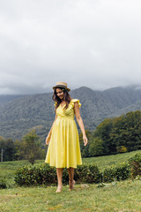Naklejka premium A young girl walk through a beautiful field. Girl in a yellow dress and a straw hat. Tea plantation, tea garden and green grass.