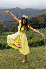 A young girl walk through a beautiful field. Girl in a yellow dress  and a straw hat. Tea plantation, tea garden and green grass.