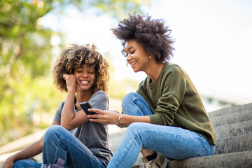 Side of two young african american women sitting on steps outside looking at mobile phone