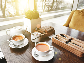 Two cups of healthy herbal tea with sea-buckthorn, ginger and honey on a textured light wooden table in a sunlit cafe with modern trendy interior. Mockup for your design.