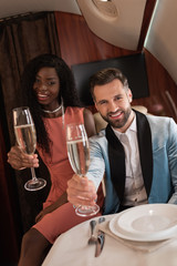 happy, elegant interracial couple holding glasses of champagne while looking at camera in private jet