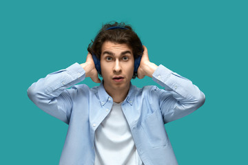 Suddenness. Portrait of a young beautiful man wearing white t-shirt and blue shirt in headphones covers ears with hands opened mouth and looking on camera