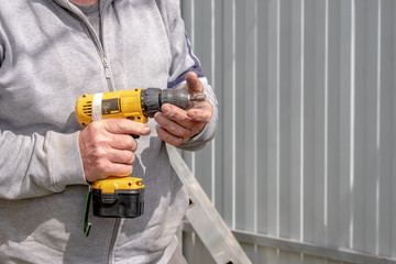 Yellow cordless screwdriver in corpus callosum male hands. Sunny day. The screwdriver is covered with scratches indicating its frequent use.