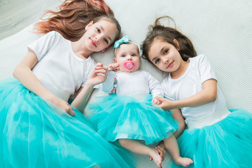 Two older sisters in beautiful ballet skirts hold their newborn sister by the arms.