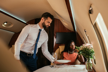 selective focus of handsome air steward serving table near elegant african american woman in plane