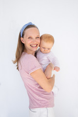 Happy positive young mom holding baby in arms, looking an camera, smiling. Young woman with little daughter isolated over outdoor white wall. Child care concept