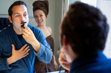 In bagno un ragazzo chiaro si fa la barba allo specchio in accappatoio, mentre la fidanzata sorride divertita 