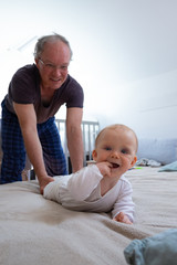 Happy grandpa playing with granddaughter in bedroom, baby biting fingers and smiling at camera. Mature man with little child in home interior. Family concept