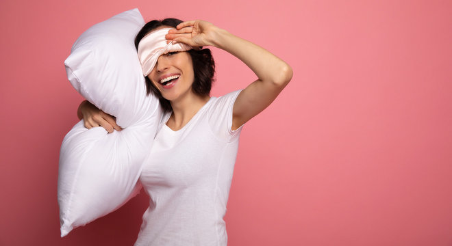 Ready For A Nap. Close-up Photo Of A Charming Woman, Who Is Fixing Her Face Mask, While Hugging A Pillow And Laughing