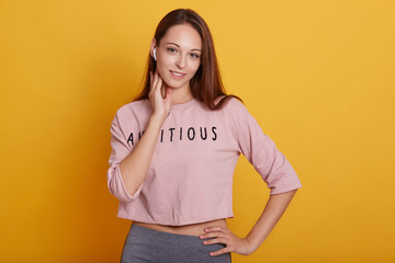 Obraz premium Picture of attractive slim female wearing rose shortened hoody and gray leggins, keeping hand neat chin, girl looking smiling directly at camera, posing isolated over yellow studio background.