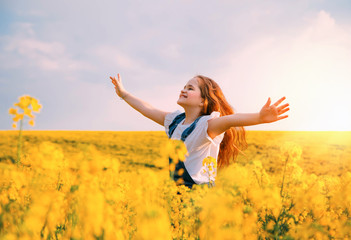 beauty young happy little girl runs, whirls at yellow flowering blooming rapeseed field. Hands raised to summer blue sky, smiling face. Red long hair fly in wind motion. Flower spring meadow at sunset