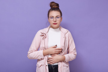 Closeup portrait of young unhappy sick female with hair bun, lady standing against lilac studio wall and keeps hands on her stomach, looks at camera, suffers from abdominal pain because of overeating