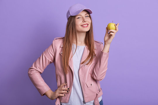 Indoor portrait of beautiful lovely teenager raising hand, holding apple, eating vitamins, being vegetarian, leading healthy lifestyle, being on diet, looking directly at camera. Food concept.