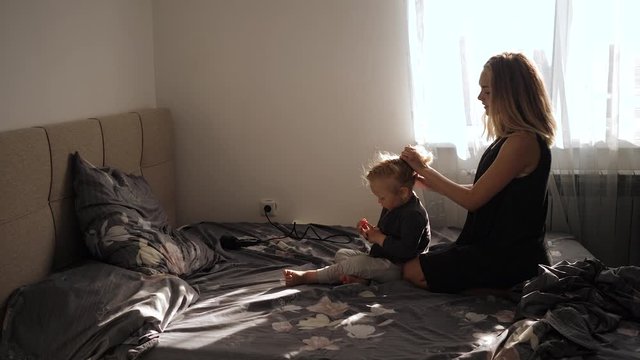 a young mother makes hairstyle little girl at home sitting on the bed