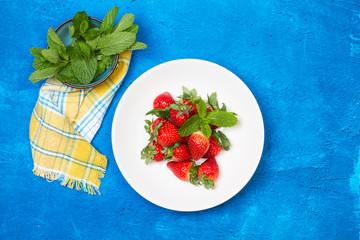 Fresas naturales y frescas dentro de un plato blanco redondo con hojas de menta sobre un fondo azul...