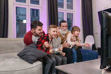 Attractive three generations of people as father, grandpa and grandchildren which sitting on the comfortable couch at home and enjoying leisure watching soccer match, shouting when team scored ball.