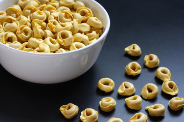 Tortellini, uncooked,  in a white bowl on a dark metallic surface