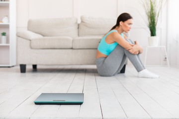 Fototapeta na wymiar Sad Woman Sitting On Floor Near Weight-Scales At Home