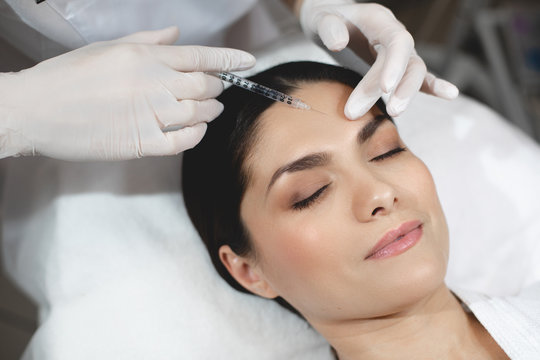 Young woman lying with closed eyes during botox injection process. Beautician in white gloves holding syringe at forehead area and input some collagen under skin.