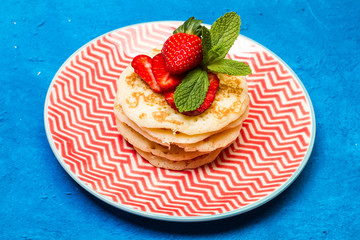 Tortitas con fresas y hojas de menta sobre un plato redondo rojo y blanco sobre un fondo azul clásico rústico. Vista superior y de frente.