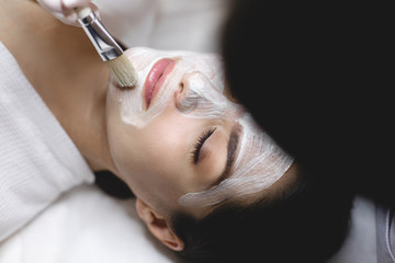 Up view of applying white facial mask process using brush. Beautician doing procedure or skin treatment for hydration. Spa time.