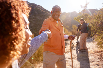 Man Helping Woman On Trail As Group Of Senior Friends Go Hiking In Countryside Together