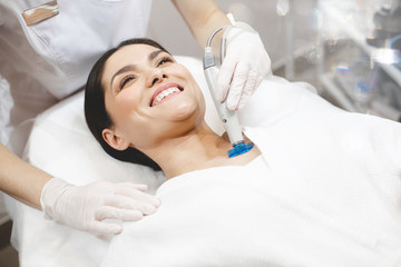 Fototapeta premium Cheerful positive young woman client lying on cosmetologist table and look up at beautician. Professional doing rejuvenation neck skin care. WElness and youthfull procedures.