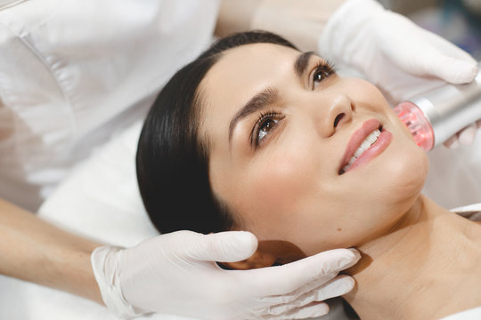 Happy beautiful positive young woman lying on cosmetologist's table. Beautician in white gloves take facial care with applying special equipment. Laser adjustment.