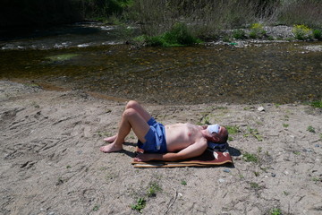 Older man with protective surgery mask sunbathing on the river bank, summer vacation in the time of coronavirus pandemic, covid 19