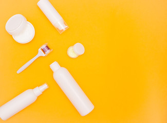 White plastic jars with cream and bottles of cosmetics on a bright background, a mesoroller and a branch with flowers. Top view, free space for text. Care cosmetics.