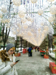 Fototapeta premium Teenager girl in glasses with a smartphone on the background of urban New Year illumination. Image with selective focus
