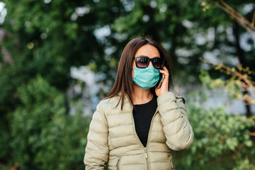 A young woman in medical mask speaks on the phone on the street against a background of greenery
