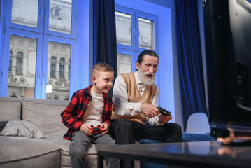 Grandfather and grandson are playing exciting video games on computer with gamepads at home.