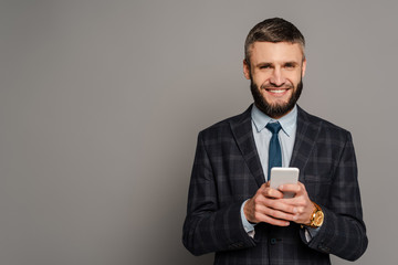 happy handsome bearded businessman in suit using smartphone on grey background