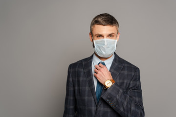 handsome businessman in medical mask adjusting tie on grey background