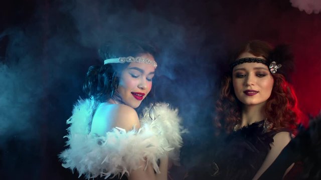 Attractive flappers women dressed in Roaring twenties time dancing on party in neon smoke room. Retro fashion, vintage lady costume