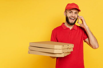 smiling bearded delivery man in red uniform with pizza boxes talking on smartphone on yellow