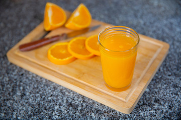Glass of fresh orange juice standing on wooden board in focus and group of orange pieces cutting with knife out-of-focus. Close-up side view. Citrus fruit, vitamin and healthy food concept
