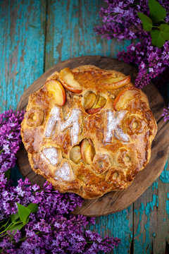 Round Homemade Pie With Apples On A Wooden Table. A Bouquet Of Lilacs. The Letters MK . Simple Pastry Made From Shortcrust Pastry And Caramelized Apple. Tart. Flour Products - Sweet Pastries.