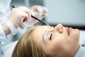 Fototapeta premium A young woman lies and master in a white uniform does eyebrow makeup in a beauty salon. The use of permanent makeup on brows. The wizard works with eyebrows. Semi-permanent makeup. Close up portrait