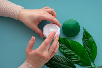 Young girls hand nearby white soft cream in jar with green leaves.Concept of eco cosmetic. Girl is touching cream as spa procedure at home.