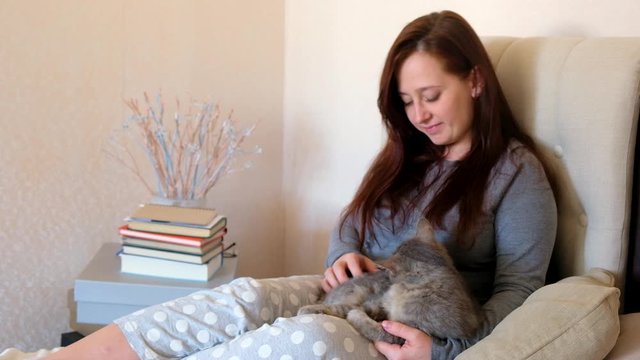 Woman stroking and cuddling grey cat on bed.  Kitten washes on girl&rsquo;s knees. Happy pet at home.