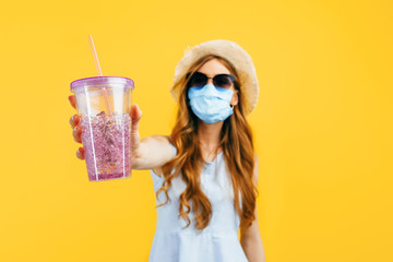 girl in a summer hat and sunglasses, wears a medical protective mask against a viral infection, holds a cocktail in her hands on a yellow background. Quarantine, coronavirus, summer